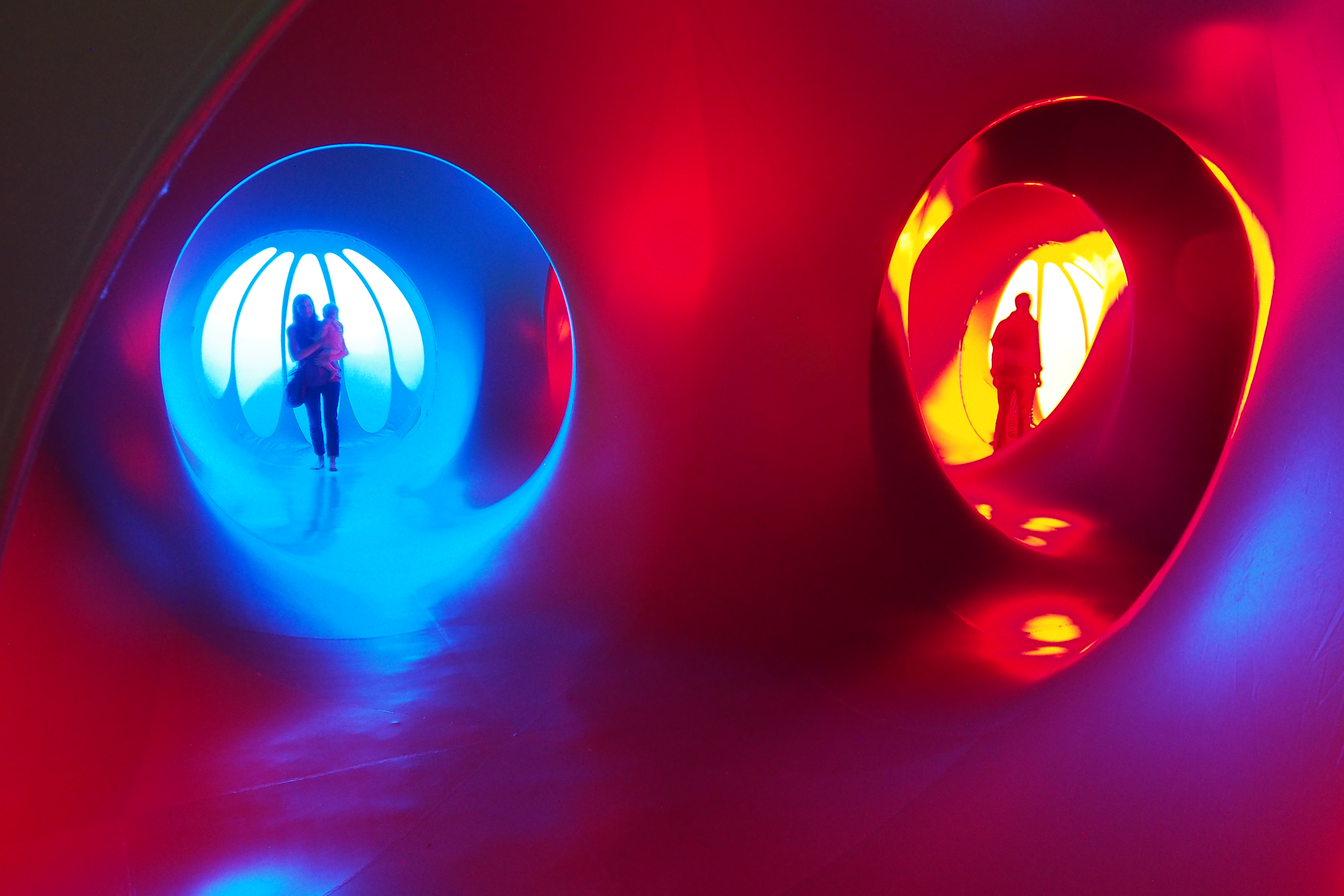 People inside a sculpture that is lit red and blue with tunnels and windows 