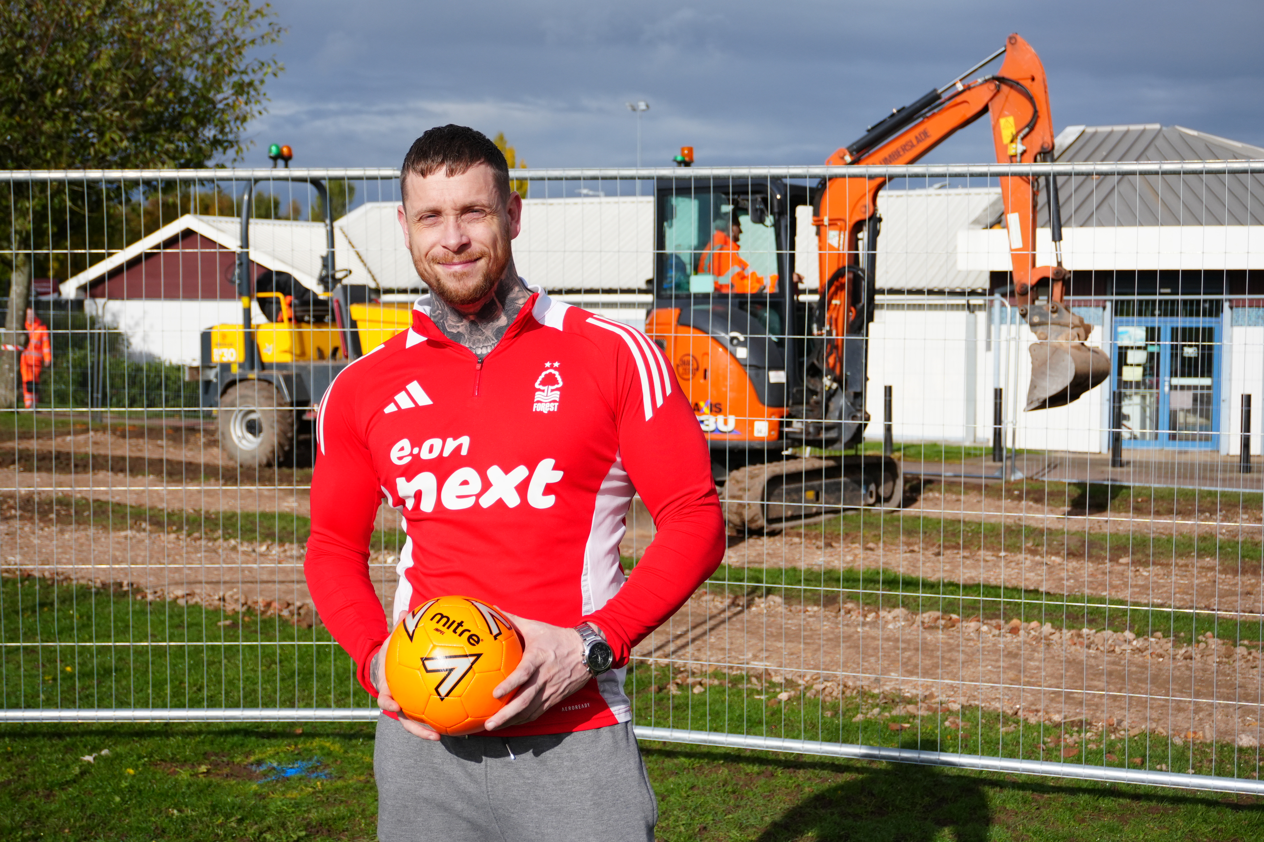 A man in a red football shirt is holding a football stood in front of the cafe building on Kingsway