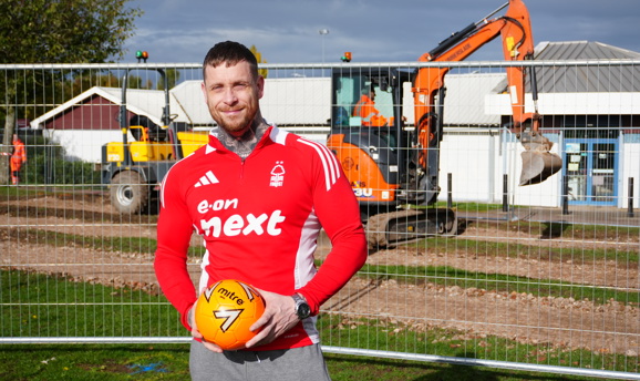 A man in a red football shirt is holding a football stood in front of the cafe building on Kingsway