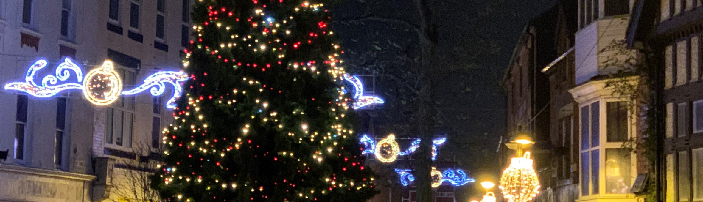 christmas tree lit up with christmas lights in the background glowing against buildings on a street