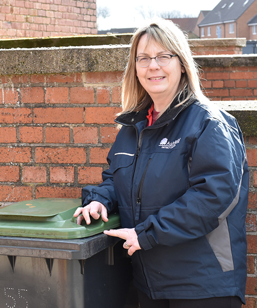 Woman looking at camera wearing a work uniform and leaning on a wheelie bin by a brick wall