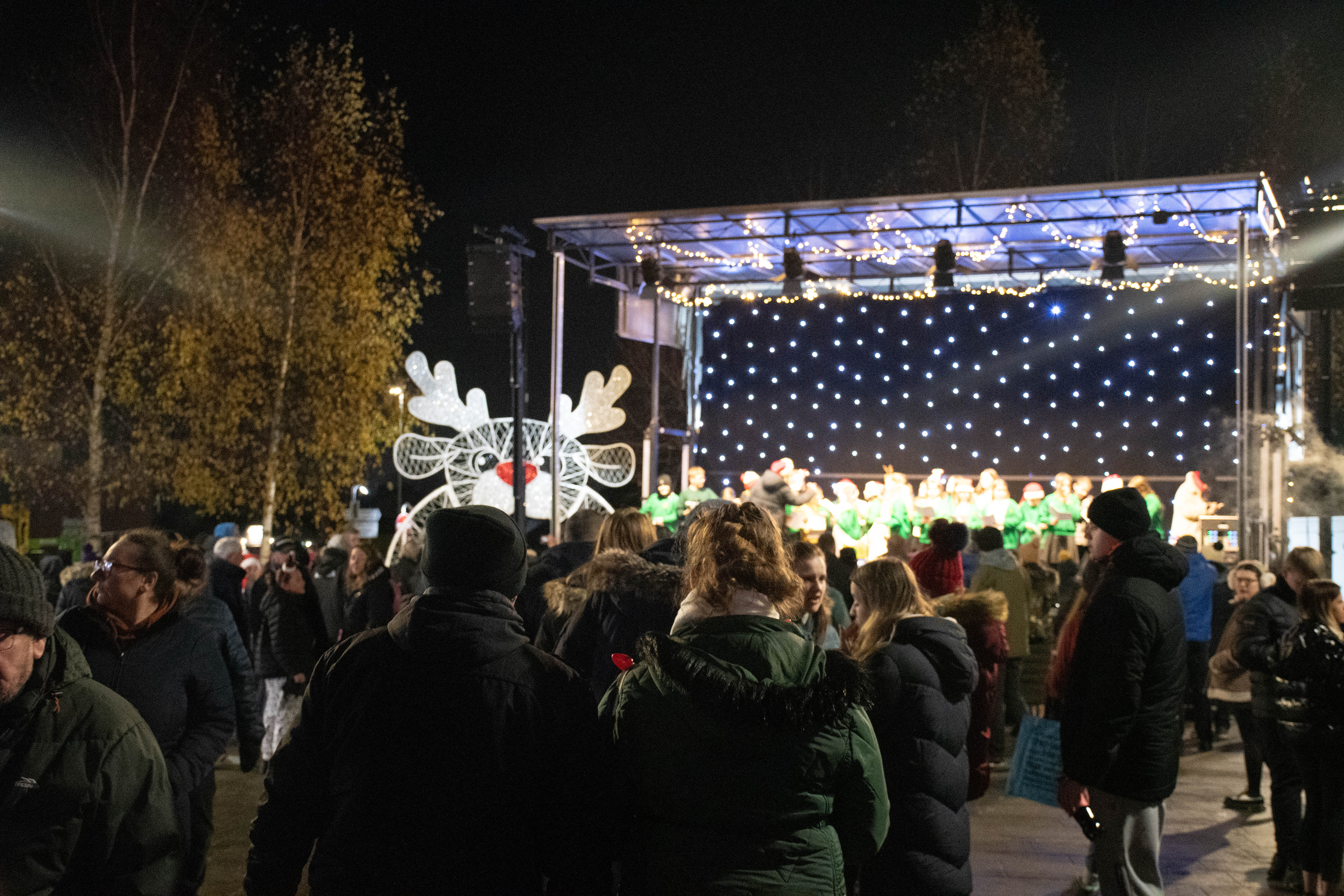 People watch a stage with a backdrop of fairy lights with a light up reindeer to the left