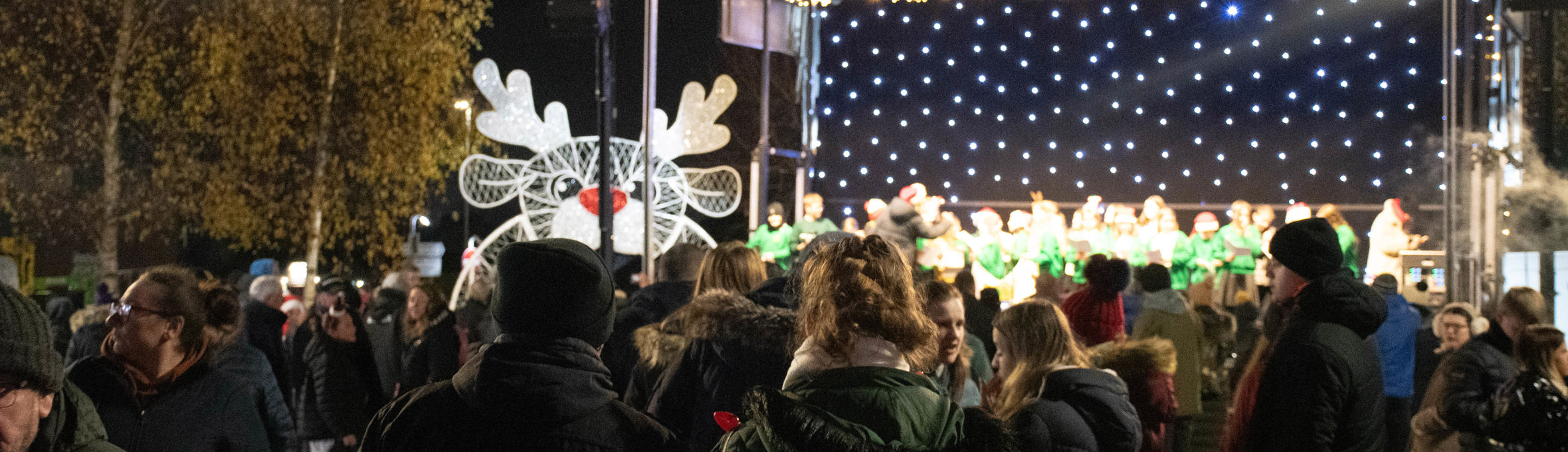 People watch a stage with a backdrop of fairy lights with a light up reindeer to the left