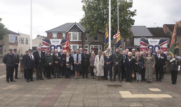 A crowd of officials and local notables beside the flags raised for Merchant Navy Day outside ADC