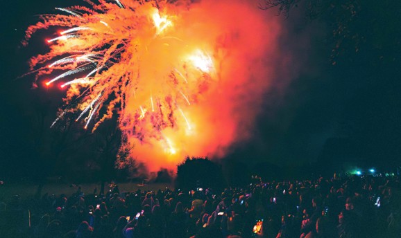 A crowd of people watching a firework display with an orange firework illuminating the crowds