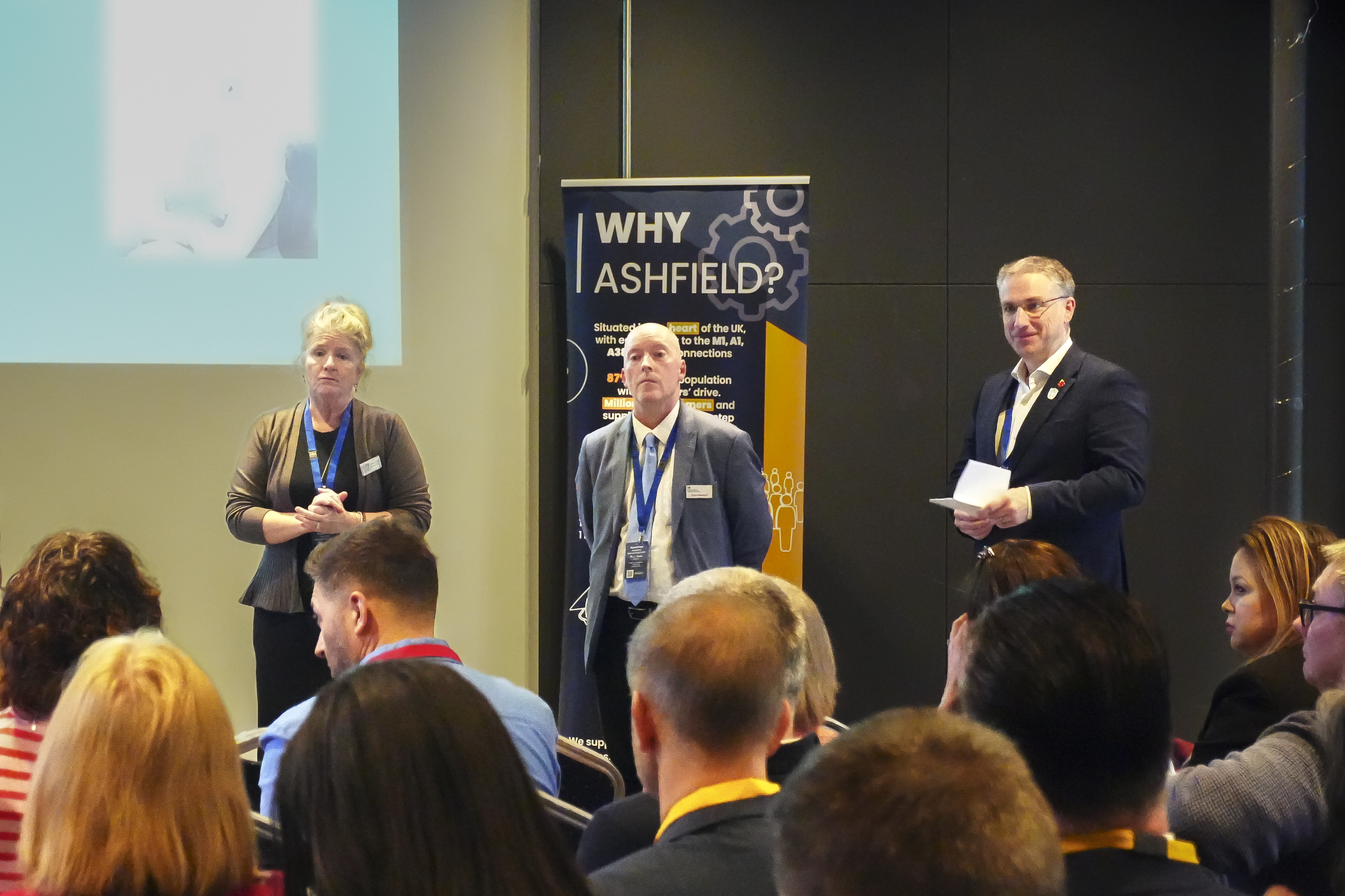 A woman and 2 men stand facing a crowd of people giving a presentation 
