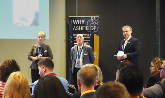 A woman and two men stand facing a crowd of people and a powerpoint presentation 