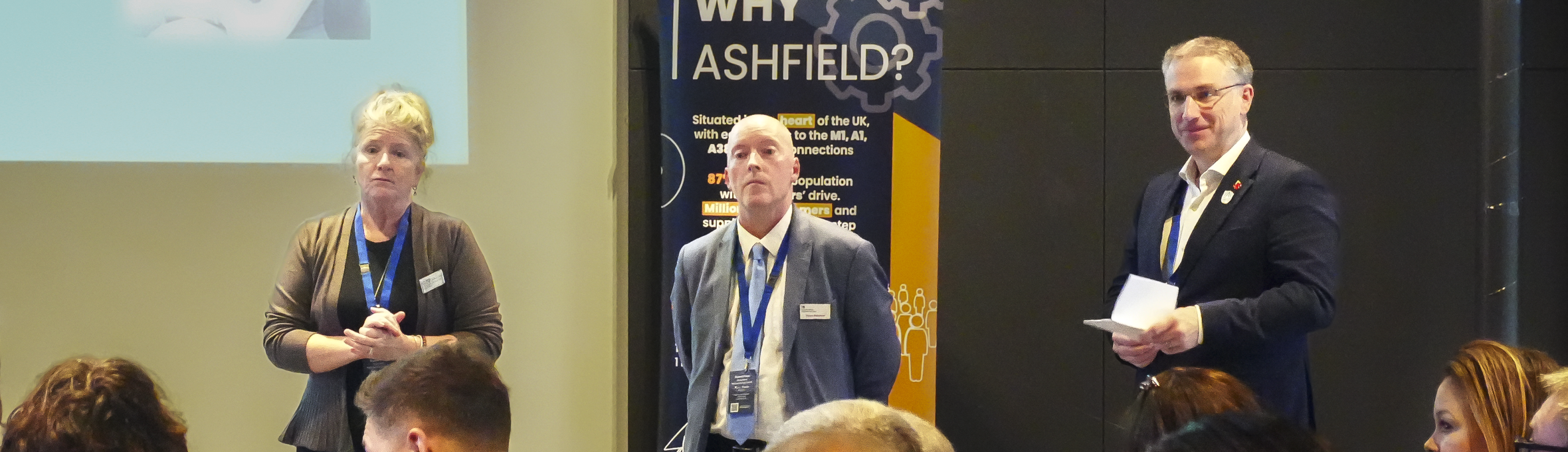 A woman and two men stand facing a crowd of people and a powerpoint presentation 