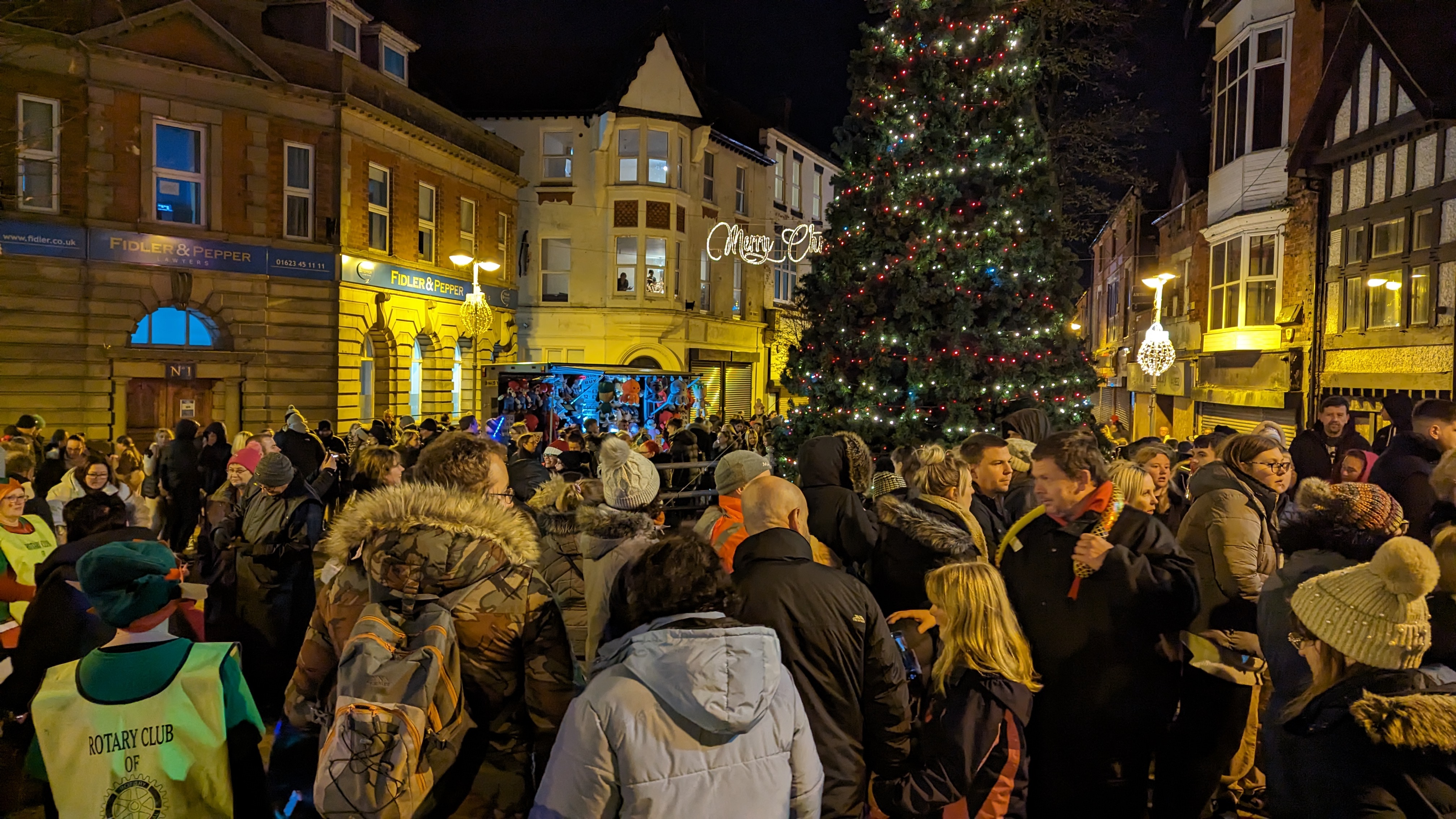 A big crowd of people are stood in front of a large Christmas tree with red and white lights on it 