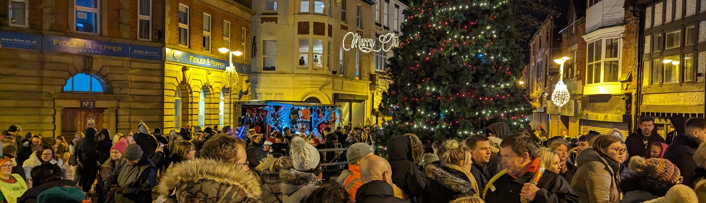 A big crowd of people are stood in front of a large Christmas tree with red and white lights on it 