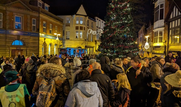 A big crowd of people are stood in front of a large Christmas tree with red and white lights on it 