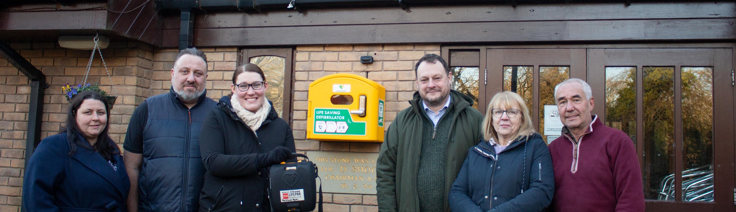 Three women and three men smile at the camera, one woman is holding a defibrillator 
