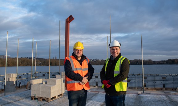 two men stand on the first floor of a building, the reservoir can be seen in the background 