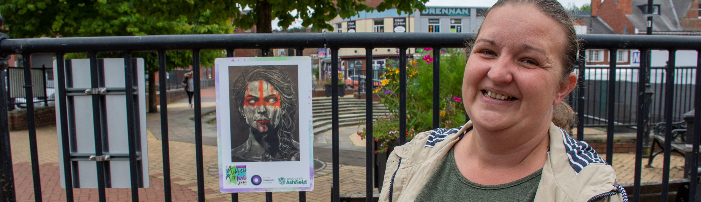 cllr sam deakin smiling next to a drawing of a woman with a red cross over her face 